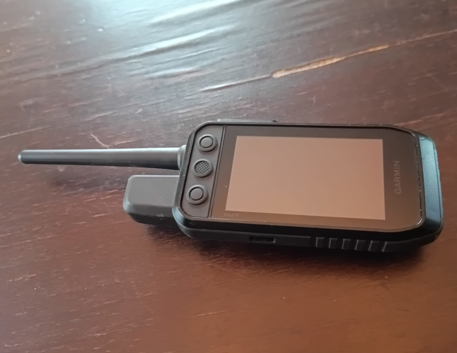 Garmin handheld GPS with attached external antenna lying on a wooden table, screen off and controls visible on the left side of the device